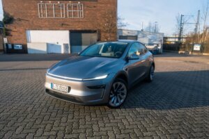 Silver Tesla Model Y parked outdoors, front three-quarter view showing the full-width light bar and 19 inch wheels
